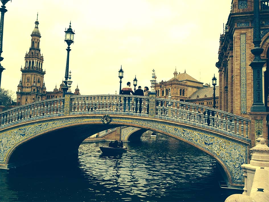 why Spain is off-limits right now. Seville Spanish plaza bridge with people on it and canoe below being rowed